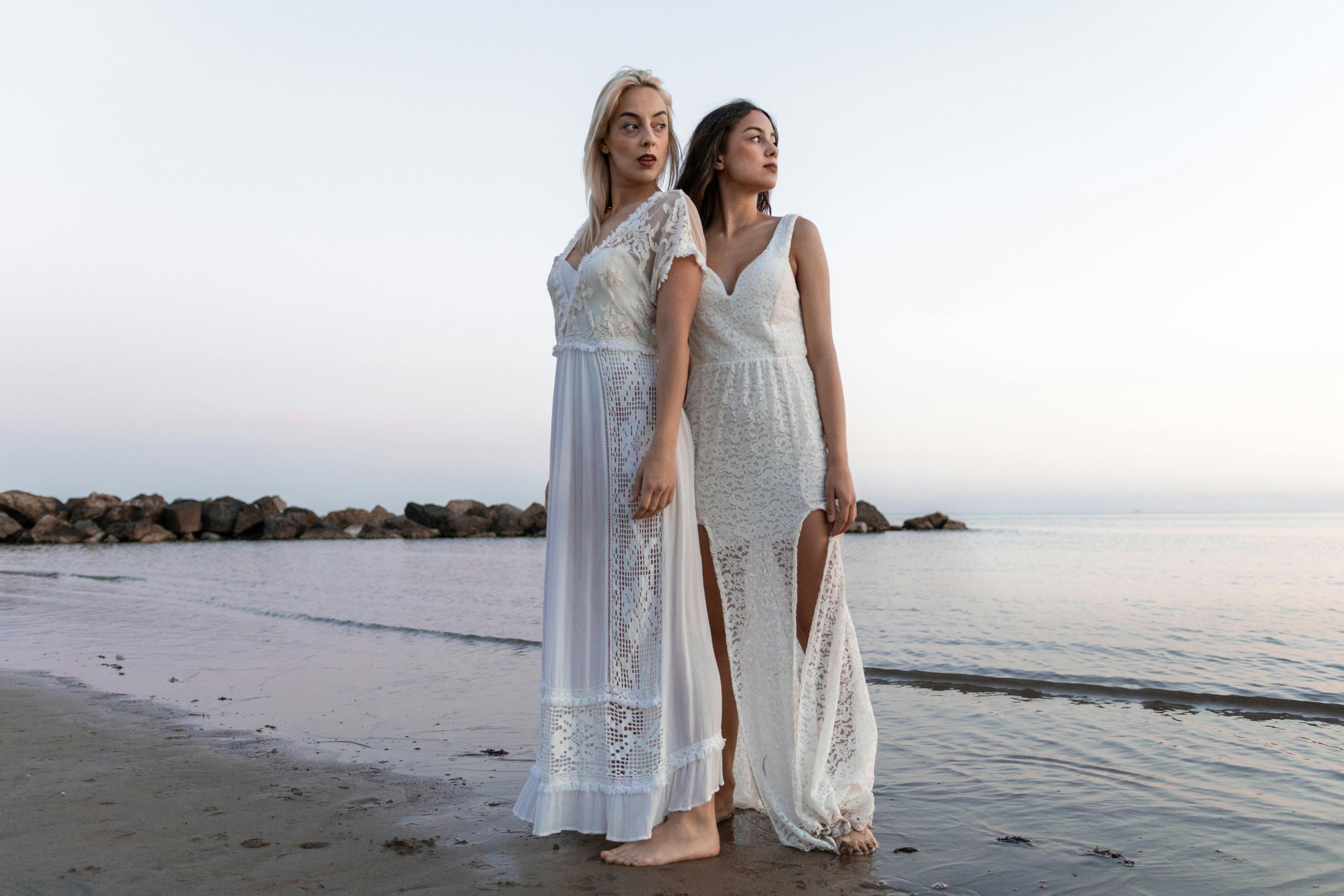 Two women in white boho style beach dresses standing on a beach with rocks and water in the background.